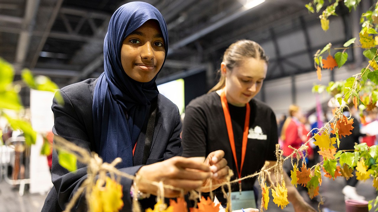 A young person at The Big Bang Fair interacts with coloured leaves at the Tree Council stand as they smile at the camera